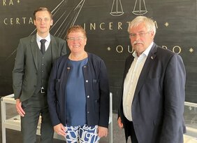 Drei formell gekleidete Personen stehen vor einer schwarzen Wand mit goldenen Schriftzügen, darunter "CERTA" und "QUIRIO", was auf einen professionellen Rahmen schließen lässt.