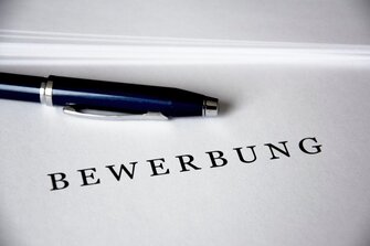 Ein schwarzer Kugelschreiber mit silbernem Clip liegt auf einem weißen Blatt Papier, auf dem in fetten schwarzen Buchstaben das deutsche Wort BEWERBUNG" steht.