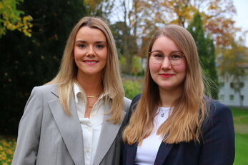 Zwei junge Frauen im Blazer stehen nebeneinander in einem Park, mit einem weißen Gebäude und Herbstbäumen im Hintergrund.