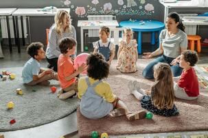 Kinder und ein Erwachsener versammeln sich in einem farbenfrohen Klassenzimmer, auf einem rosa Teppich sitzend und umgeben von buntem Spielzeug und Lernmaterial.